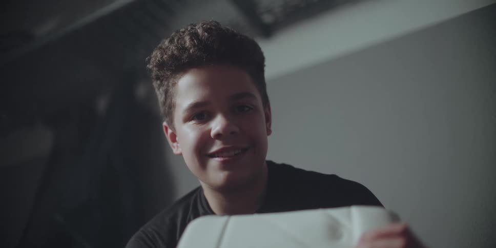 A young hockey goalie with curly hair smiles while looking directly at the camera in a locker room