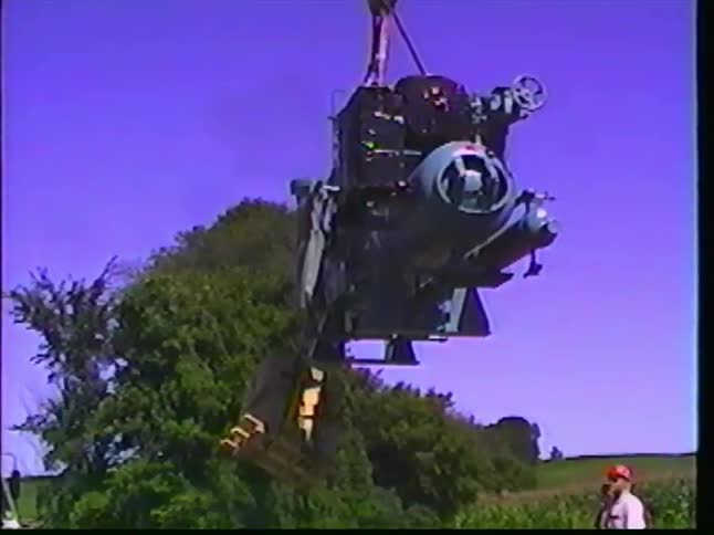 Archival footage captures workers carefully using a large crane to lift and move heavy industrial machinery from a truck at a worksite