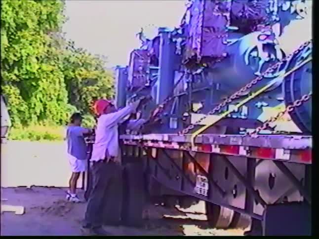 Retro VHS footage captures two workers carefully securing large industrial machinery onto a flatbed truck with chains