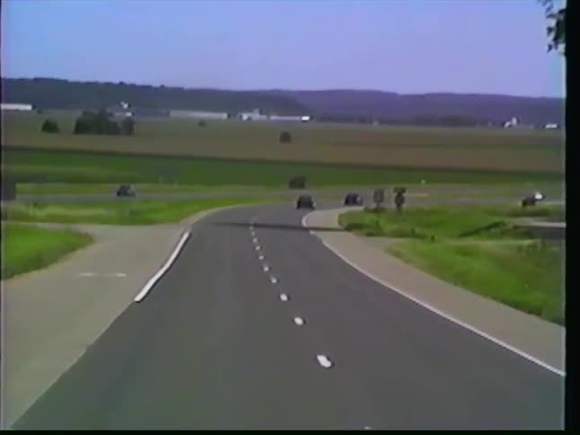 Vintage VHS footage captures a truck driver's point of view while navigating a curving highway ramp through the rural American heartland on a sunny day