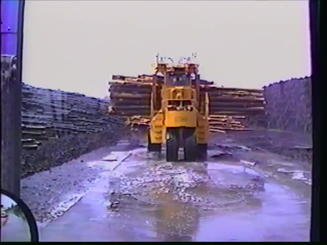 A large yellow log loader carries a heavy load of timber as it drives through a muddy, wet path at a lumberyard