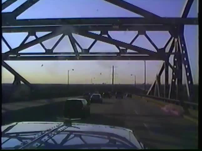A vintage VHS point-of-view shot from a vehicle driving across a steel truss bridge, passing an industrial port town
