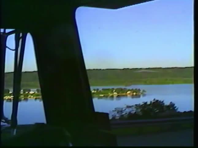 A scenic view of a wide river and forested hills is captured from the window of a moving vehicle in this vintage footage