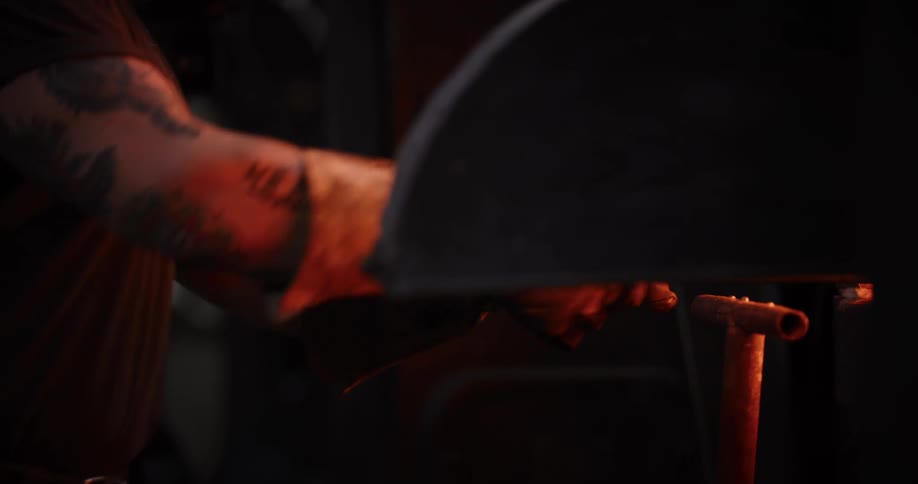 A blacksmith with tattooed arms wearing protective gloves removes a white-hot piece of metal from a furnace in a dark forge