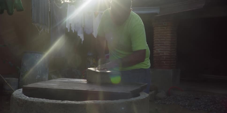 Sun flares behind a man in a green shirt as he sharpens a blade on a large stone in a rustic yard