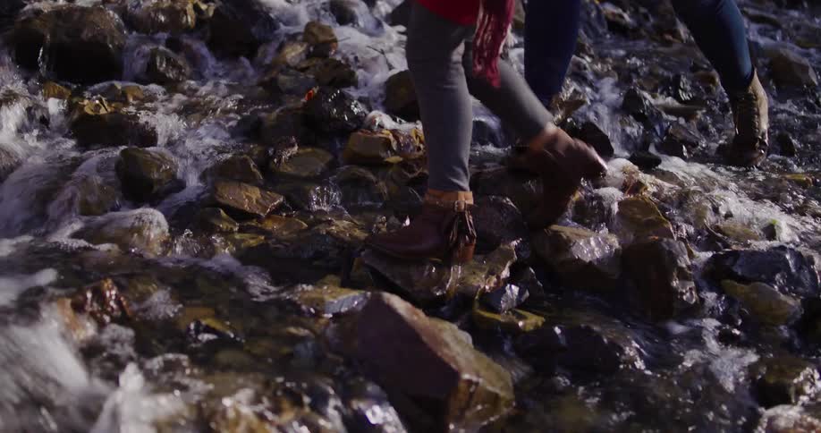 Carefree mother and daughter hold hands and giggle as they walk across rocks in a rushing stream in slow motion