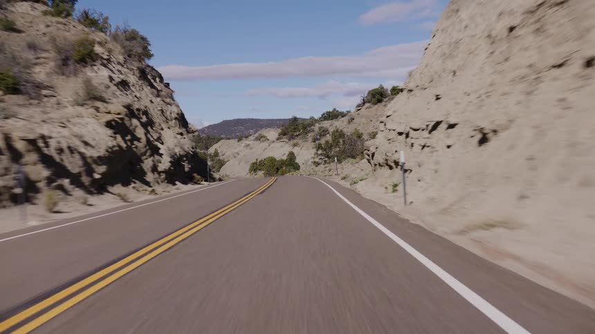 A point-of-view shot from a vehicle driving along a winding, empty desert highway through rocky hills on a sunny day