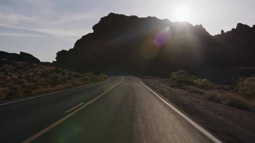 A slow-motion driving shot travels down a winding desert road as the sun flares over a large, silhouetted rock formation