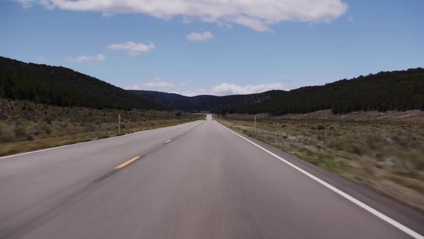 A point-of-view shot driving along an empty, scenic highway through a rural valley with rolling hills under a blue sky