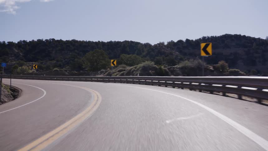 A point-of-view shot of a car driving along a winding paved road through a rocky, arid mountain landscape on a sunny day