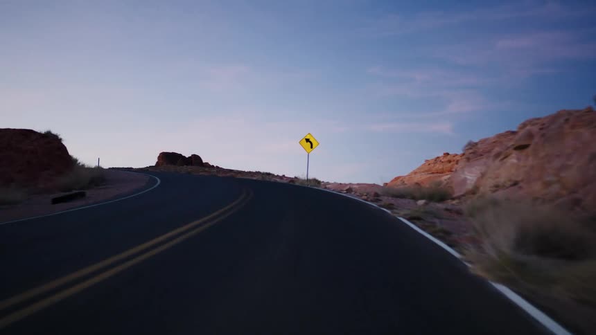 A smooth POV drive on a winding desert highway through red rock formations at dusk approaches a yellow curve sign