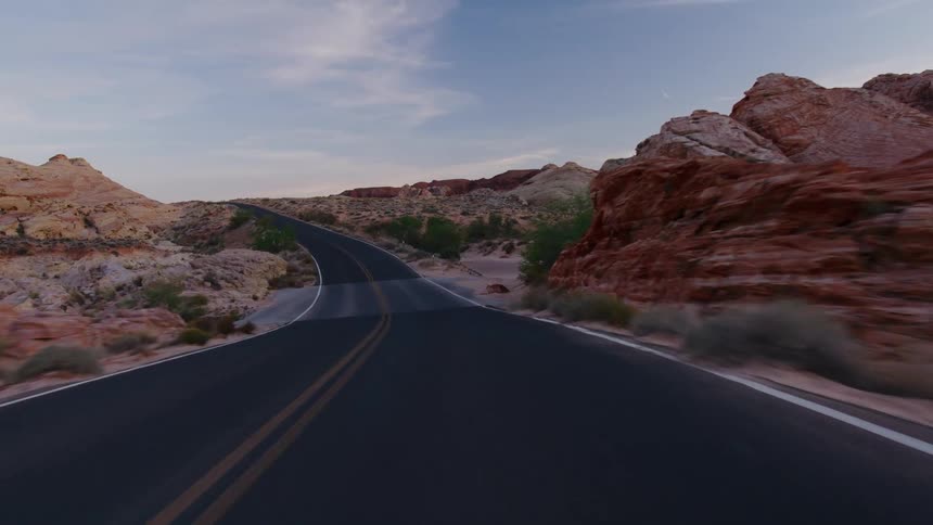 A scenic drive on an empty, winding road through a desert canyon landscape with colorful rock formations at twilight