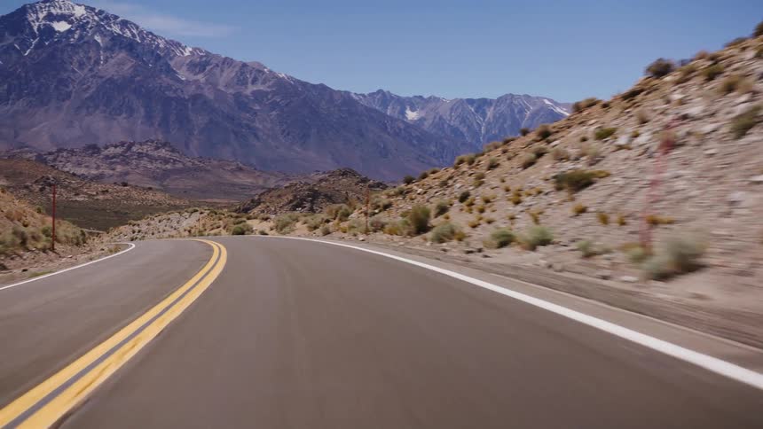 POV driving on a winding desert highway reveals majestic snow-capped mountains under a clear blue sky
