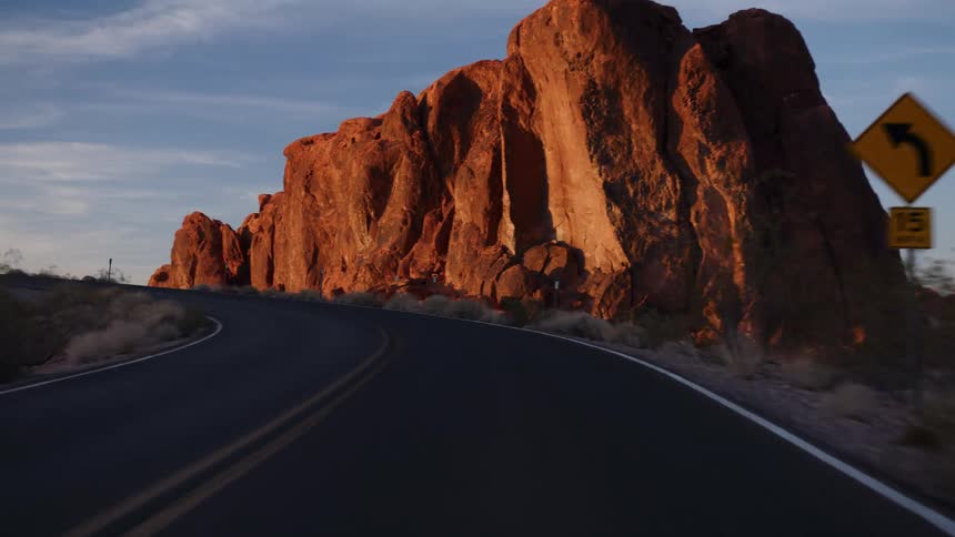 A scenic point-of-view drive along a winding desert road passes by majestic red rock formations illuminated by the golden light of sunrise