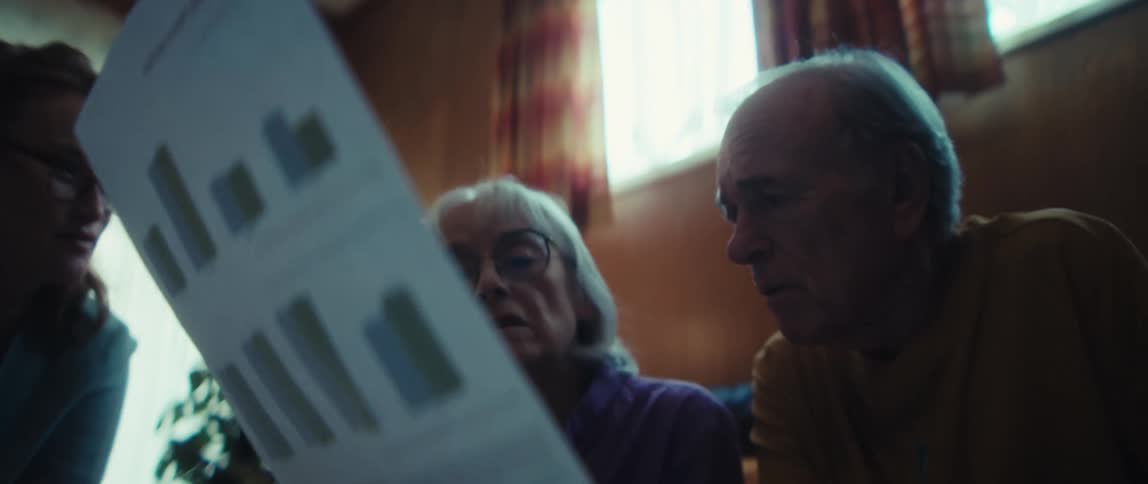 A rack focus shot reveals a worried elderly couple as they review a document with bar graphs in their home