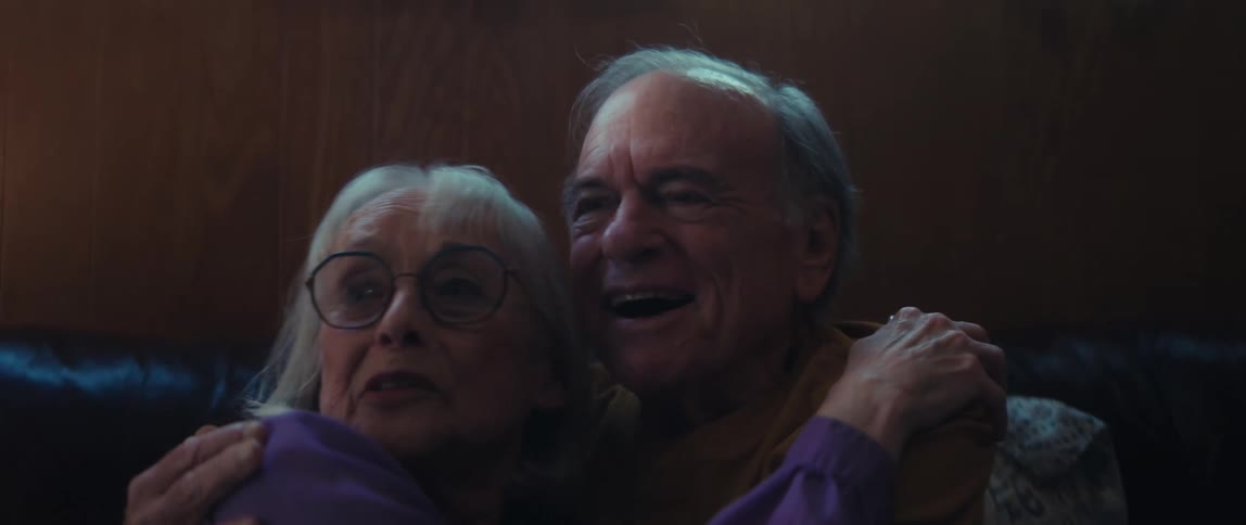 An expressive elderly couple reacts with both fear and amusement while watching a movie together on the couch