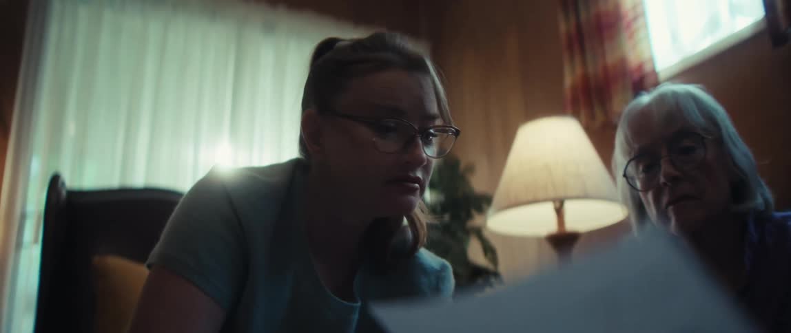 A young woman with glasses carefully explains a document with bar charts to an older woman in a dimly lit room