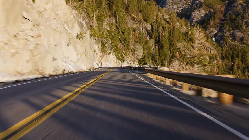 A point-of-view shot captures a smooth drive on a winding road through a rocky mountain pass with pine trees and lingering snow