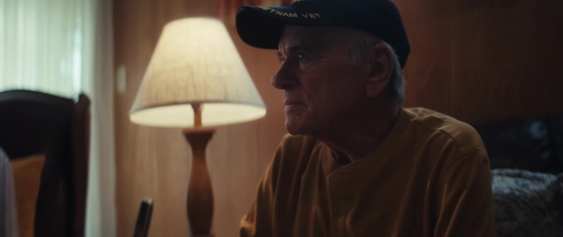A thoughtful elderly Vietnam veteran in a cap sits in a dimly lit room, reflecting