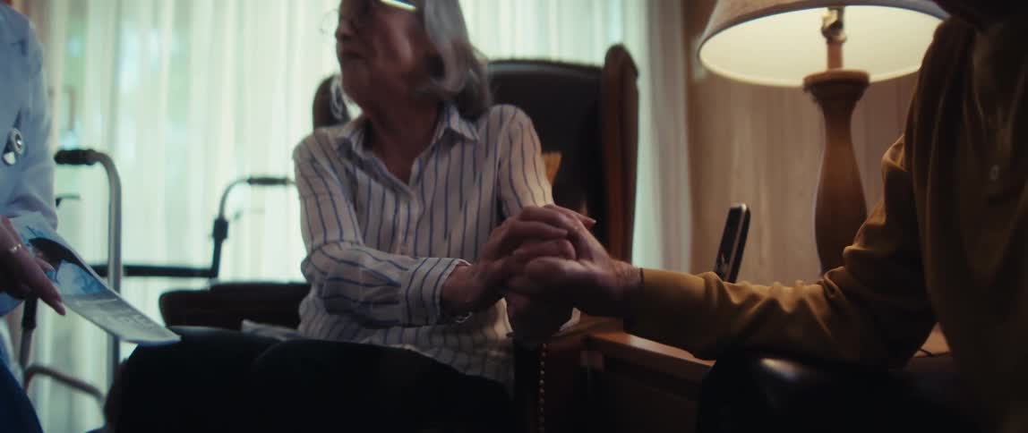 In a tender moment, an elderly husband holds his wife's hand for support during a home visit from her doctor