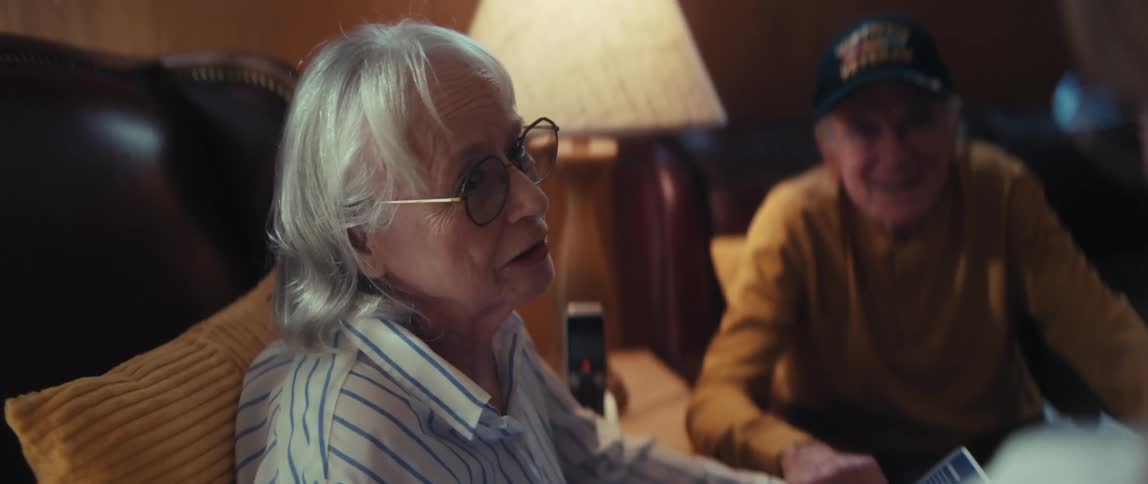 An elderly Vietnam veteran smiles and listens as his wife animatedly tells a story to a guest in their living room