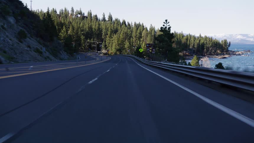 A POV shot of a scenic drive along a winding road next to a large blue lake surrounded by pine forests and mountains