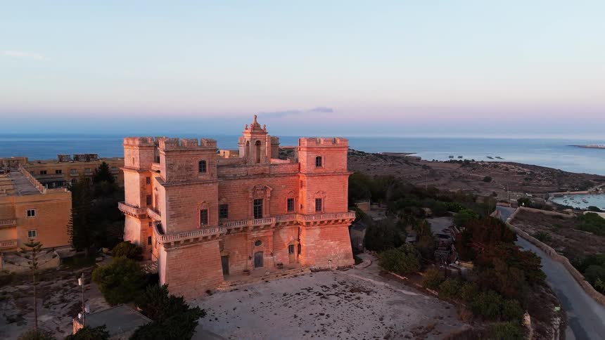 An aerial orbit reveals the historic Selmun Palace in Malta, glowing in the warm light of sunset with the sea in the background