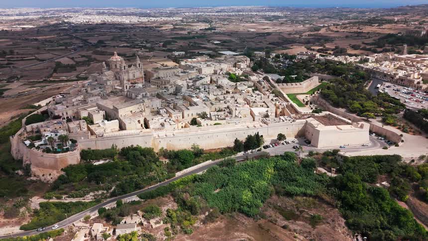 A wide, static aerial view captures the ancient fortified city of Mdina in Malta, perched on a hill above the surrounding countryside