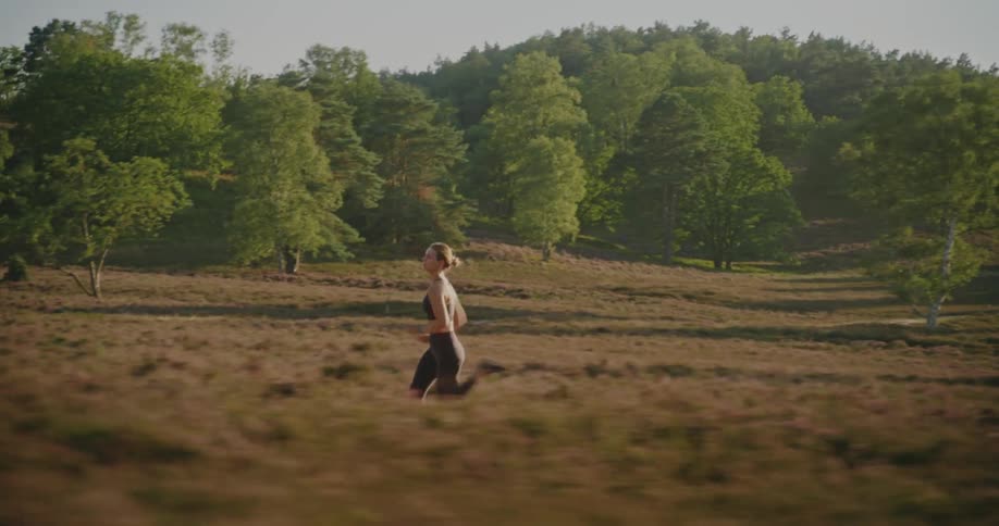 Slow motion tracking shot follows a fit woman running through a scenic field with a forest in the background during golden hour