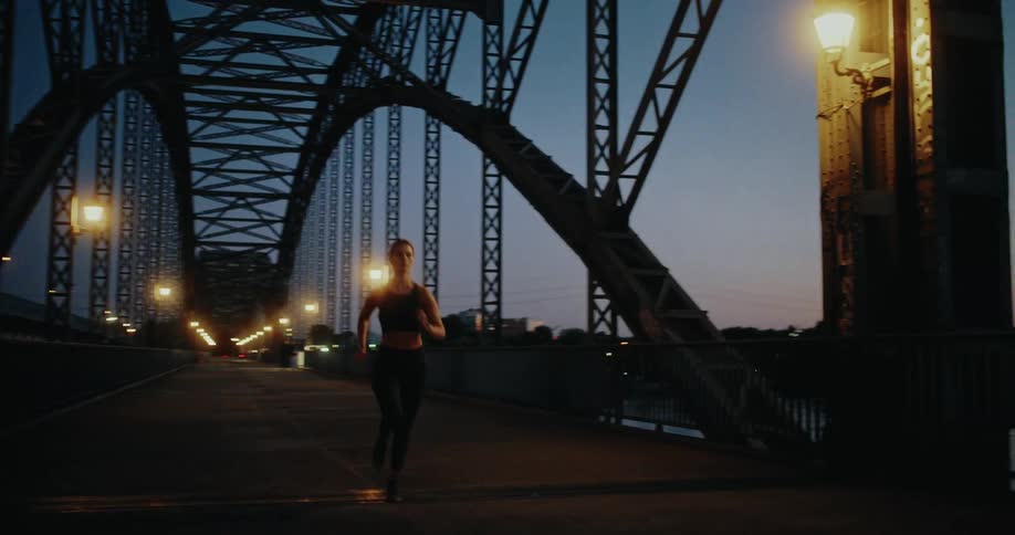A fit woman runs across a large illuminated city bridge at dusk