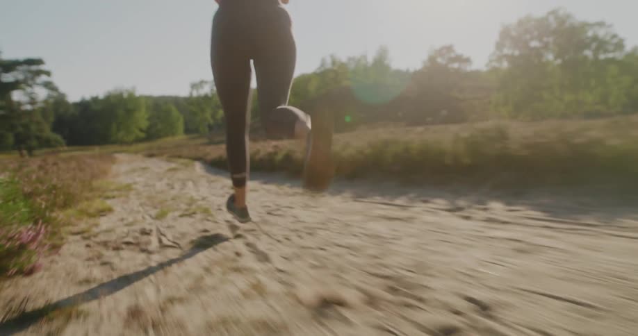 Dynamic slow-motion shot follows a woman running on a sandy trail through a sunny, natural landscape