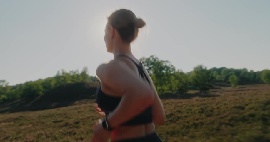 Slow motion medium shot from behind of an athletic woman running through a beautiful sun-drenched field in nature