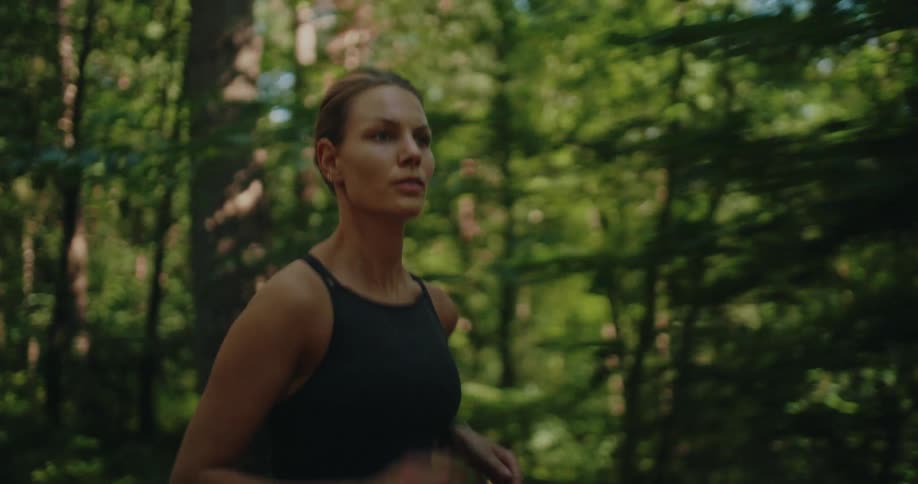 A close-up of a determined athletic woman jogging through a sunlit forest
