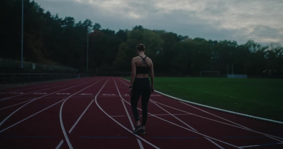 From behind, a female athlete in black sportswear walks slowly along a running track to cool down at dusk