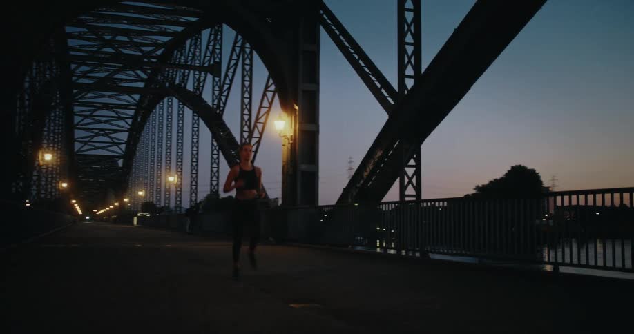 A determined athletic woman jogs in slow motion across a large industrial bridge at dusk