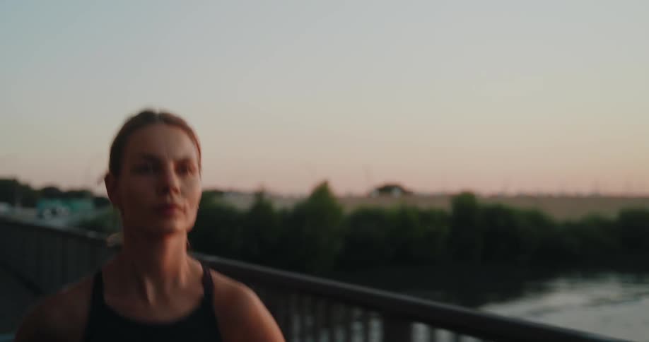 An athletic woman in slow motion runs across a bridge with determination at sunset