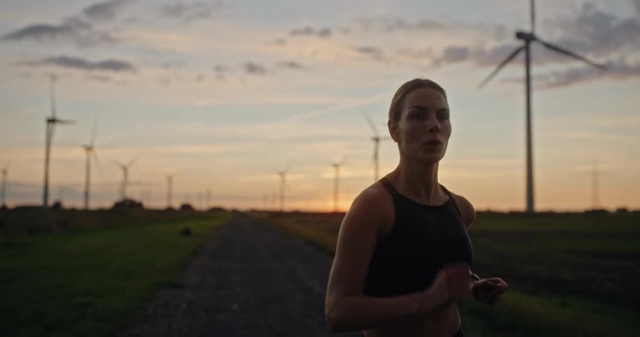 An athletic woman runs in slow motion on a rural road, with a field of wind turbines silhouetted against the beautiful sunset sky
