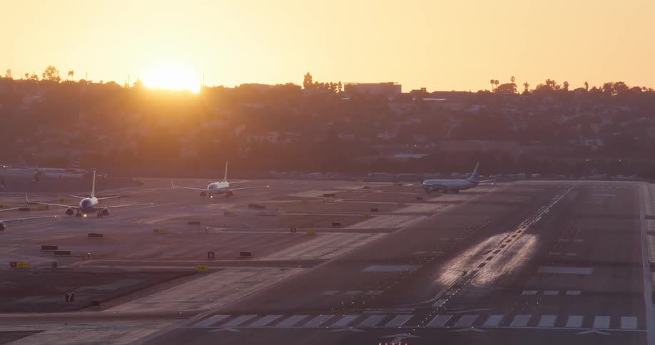 Several airplanes taxi on the runway at a busy airport as the sun sets in the distance