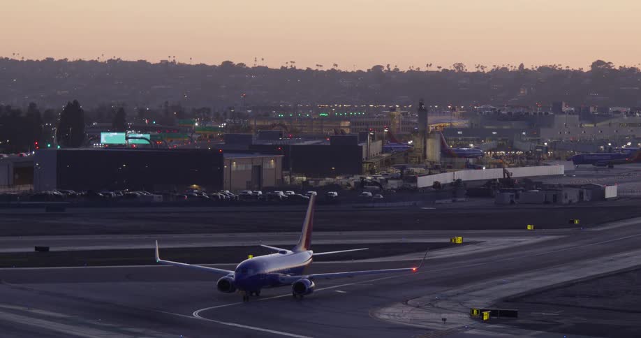 A stationary passenger airplane waits on the runway of a busy urban airport as the evening lights begin to turn on at dusk