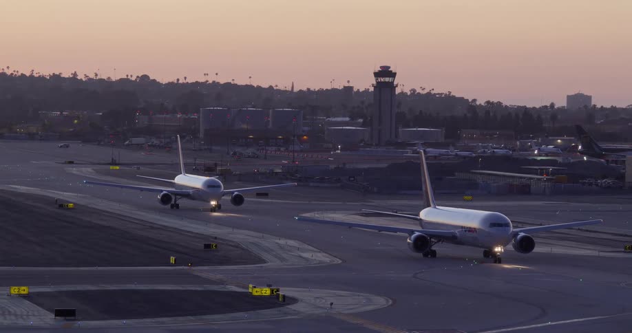 Two commercial jets with lights on taxi along the runway at a busy airport at dusk