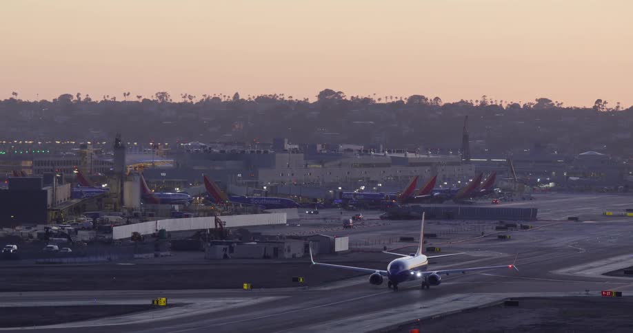 A passenger airplane with its lights on taxis down the runway at a busy airport terminal during a colorful sunset