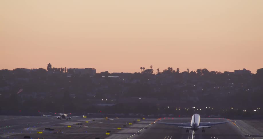 A passenger airplane accelerates down a brightly lit runway and takes off into the dusky sky with a cityscape in the background