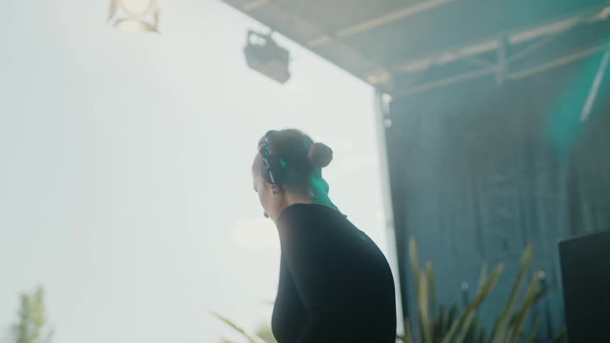 A female DJ seen from behind prepares her mix, putting on headphones to get into the groove at a daytime festival