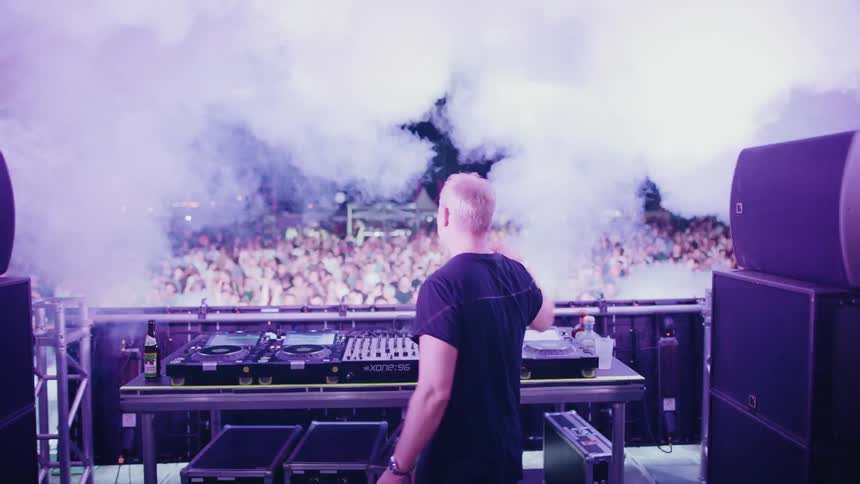 A DJ hypes up a massive crowd from the stage at a vibrant nighttime music festival, complete with strobe lights and smoke machines