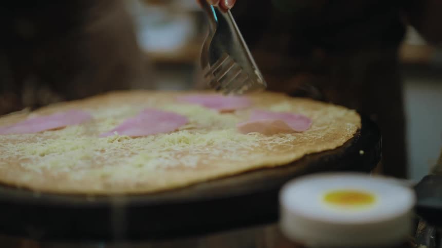 A chef carefully places slices of ham onto a savory, cheesy crêpe cooking on a hot griddle in slow motion