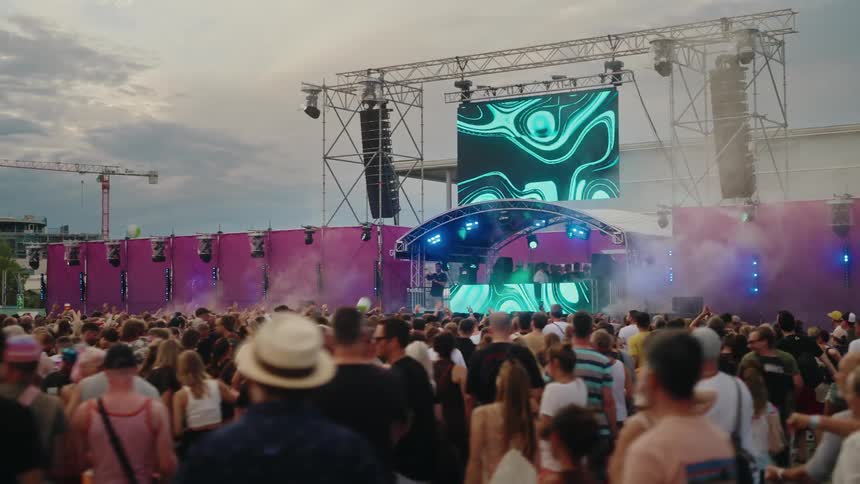 A massive crowd dances and vibes to the electronic music played by a DJ on a large outdoor festival stage with vibrant visuals and smoke effects