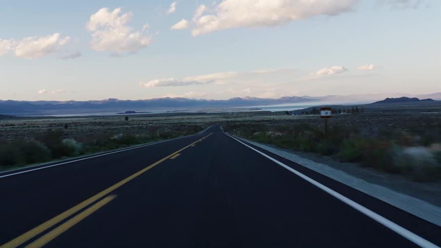 A smooth point-of-view shot driving down an empty highway through a desert landscape towards a tranquil lake and mountains at sunset