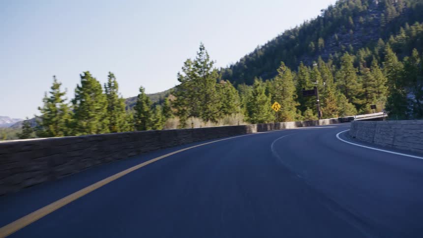A smooth point-of-view shot emerges from a dark highway tunnel onto a sunlit, winding mountain road in a forest setting