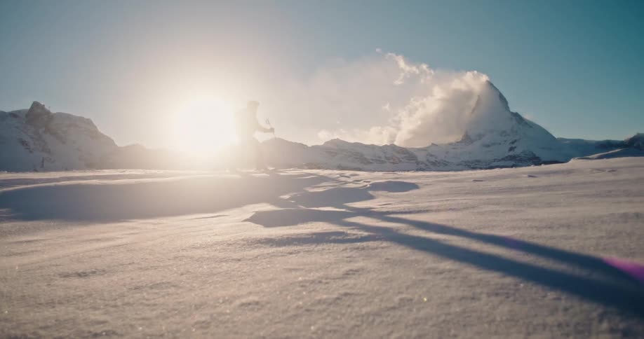 A mountaineer hikes across a vast, sun-drenched snowfield in slow motion, with the majestic Matterhorn peak in the background