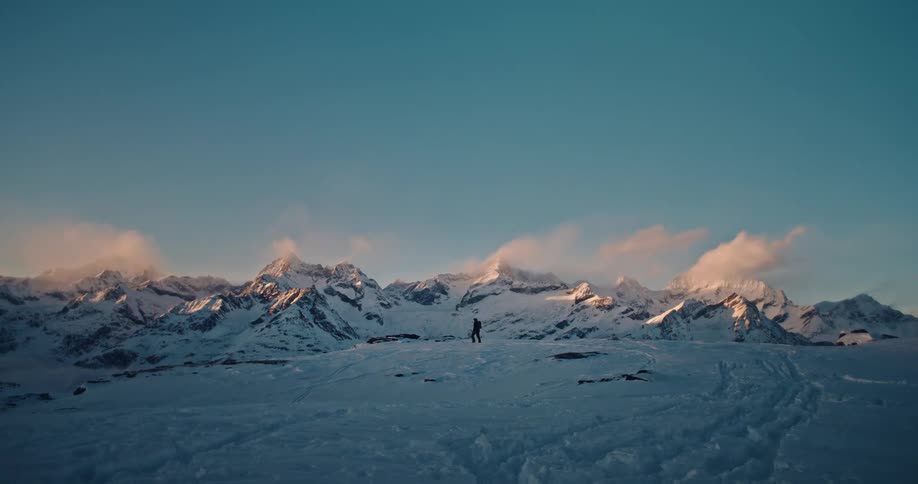A solitary hiker treks across a vast snowy landscape with a breathtaking mountain range illuminated by the sunrise in the background